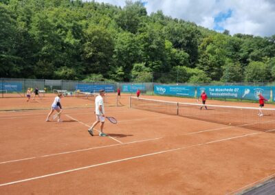 Zwei Tennisgruppen spielen auf einem roten Sandplatz mit grüner Umgebung und Werbebannern im Hintergrund.