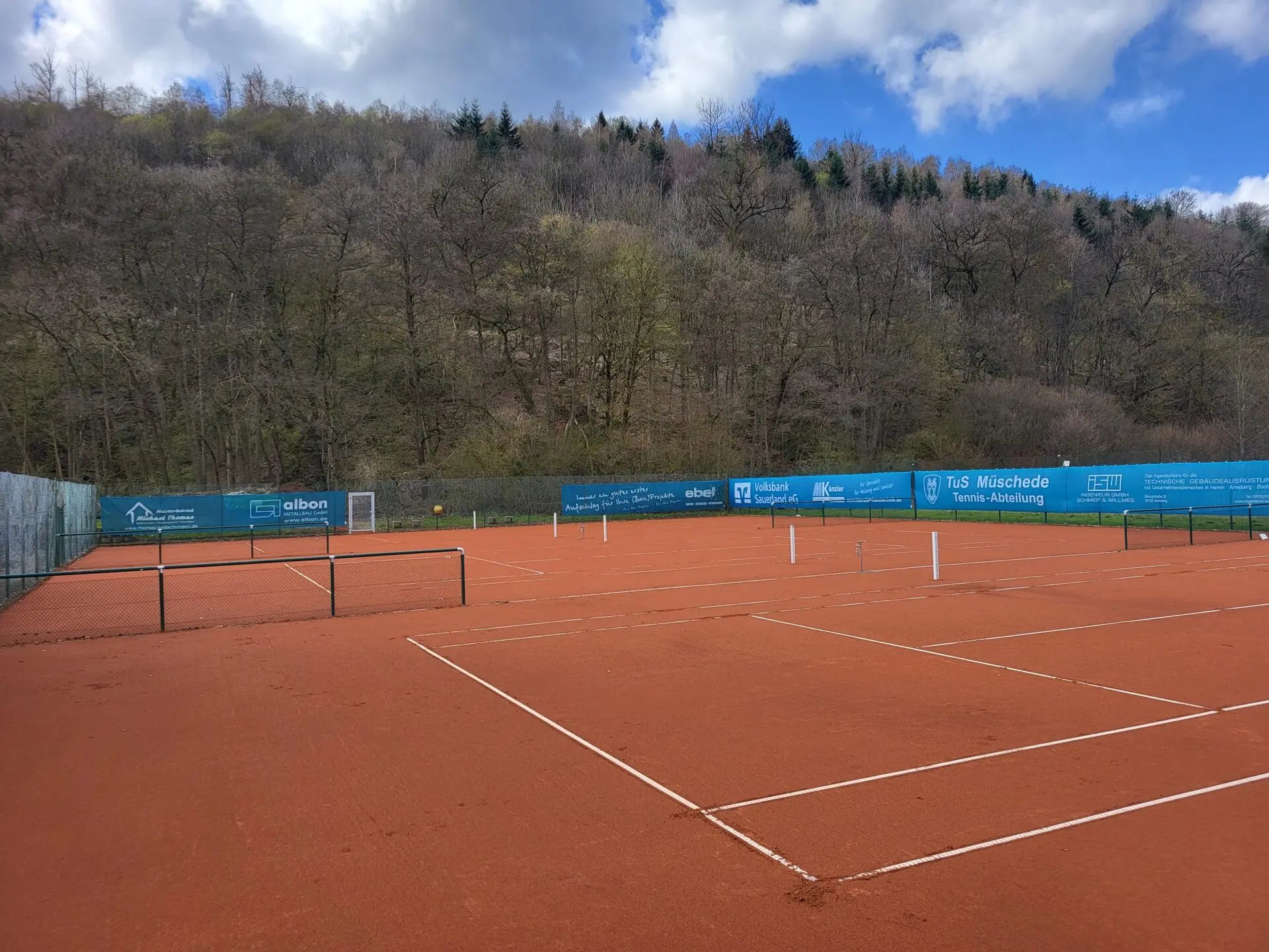 Tennisplätze aus rotem Sand mit blauen Werbebannern der TuS Müschede Tennis-Abteilung im Hintergrund, umgeben von Baumhügeln und einem klaren Himmel.
