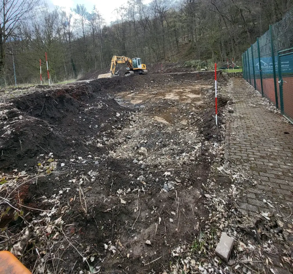 Gelber Bagger auf einer Baugrube mit Kies, rot-weiße Markierungspfosten am Rand, Zaun rechts und dunkle Erde im Hintergrund
