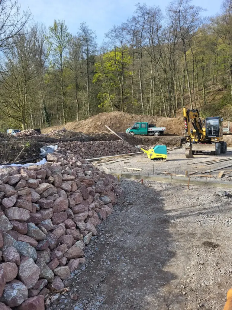 Baugelände im Wald: gelber Bagger und Baufahrzeug auf Schotterweg, Erdhügel und Steinwälle, umgeben von grünen Bäumen unter blauem Himmel.