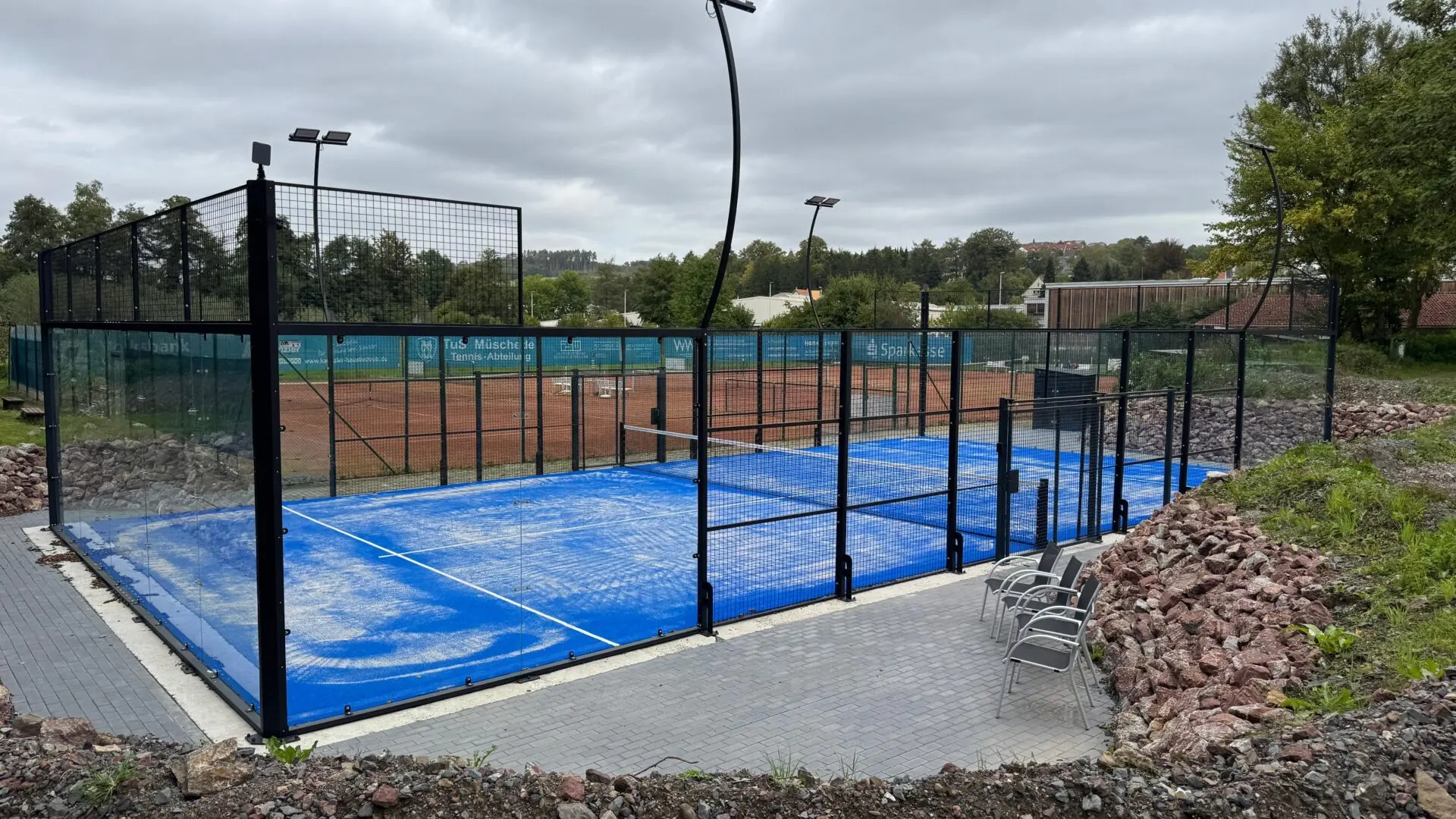 28009 Die blaue Padelanlage des TuS Müschede mit Glaswänden, Netz und Beleuchtung liegt neben Tennisplätzen unter bewölktem Himmel.