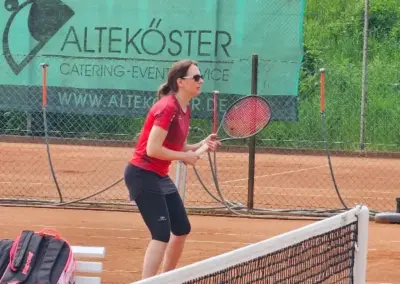 Rote Tennissportlerin mit Schläger steht am Netz auf rotem Sand, im Hintergrund ein grünes Banner mit Aufschrift „ALTEKÖSTER“