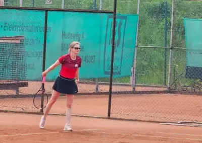 Tennisspielerin im roten Shirt und schwarzem Rock schlägt mit dem Schläger auf dem Sandplatz, Platz 3, mit grünem Zaunbanner und Wald im Hintergrund.
