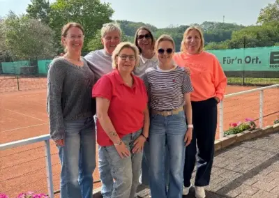 Auf dem Tennisplatz stehen sechs Vereinsmitglieder in roten, orangefarbenen und gestreiften Shirts, mit Sonnenbrillen und strahlendem Lächeln.